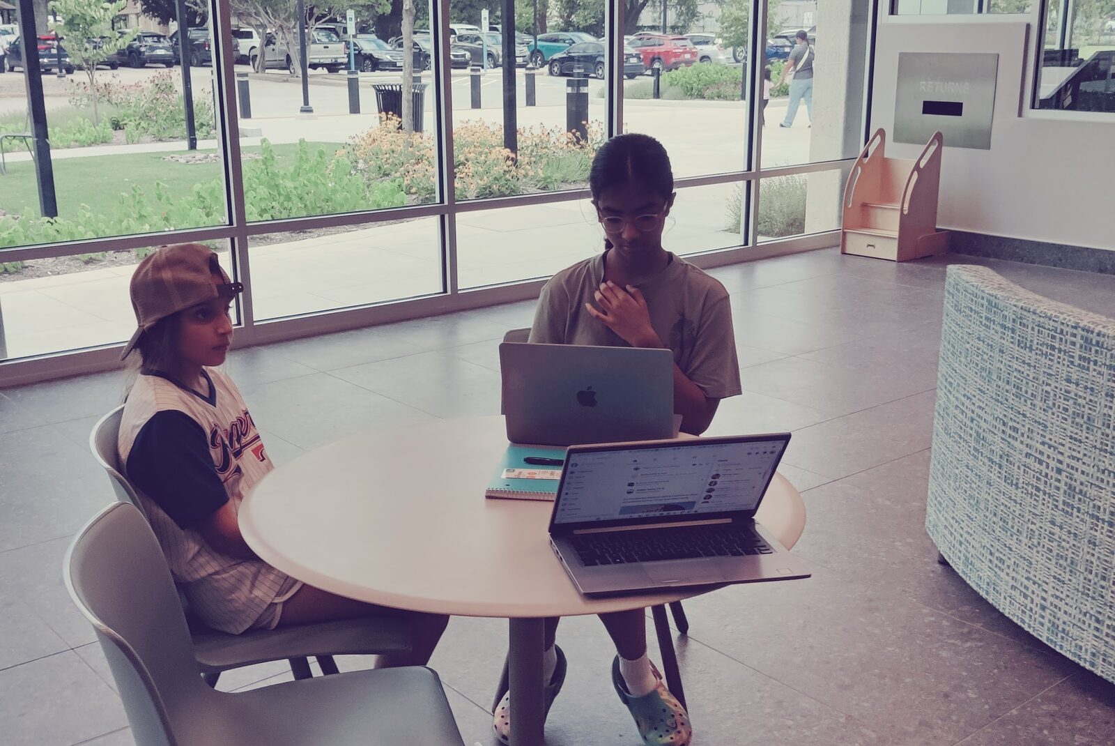 Two students studying on laptops at a table beside the library's floor-to-ceiling windows.